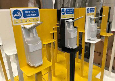 Yellow, black, and white powder coated hand sanitiser stands with mounted dispensers in workshop