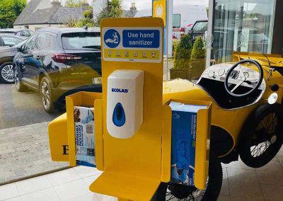 Yellow powder coated hand sanitiser stand with mounted dispenser and brochure holders in showroom