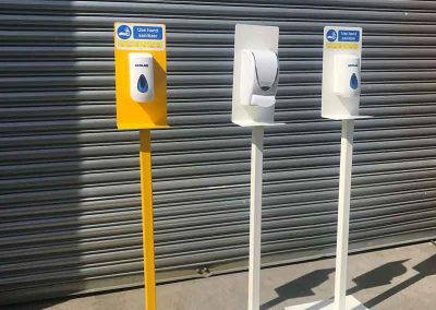 Yellow and white powder coated hand sanitiser stands with dispensers displayed outdoors